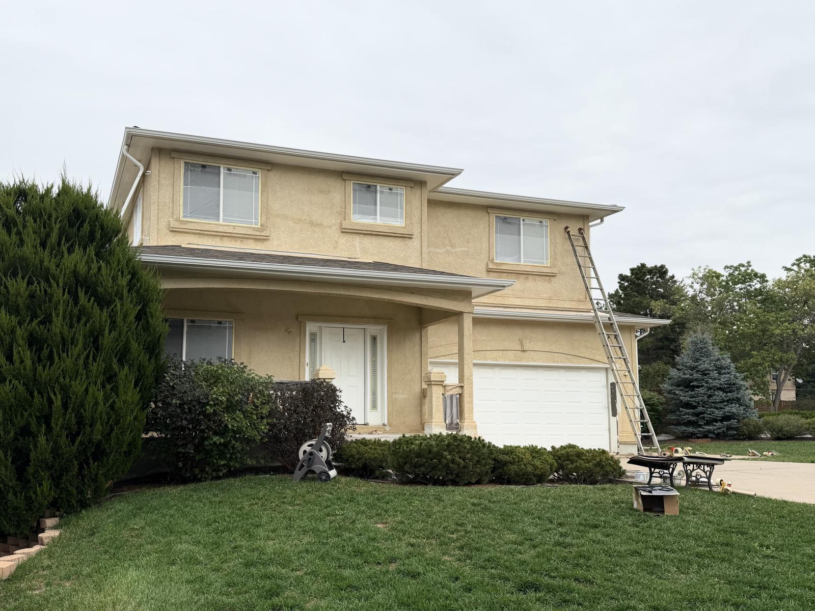 Before: two-story stucco home, front elevation, faded yellow with white trim, ladders up